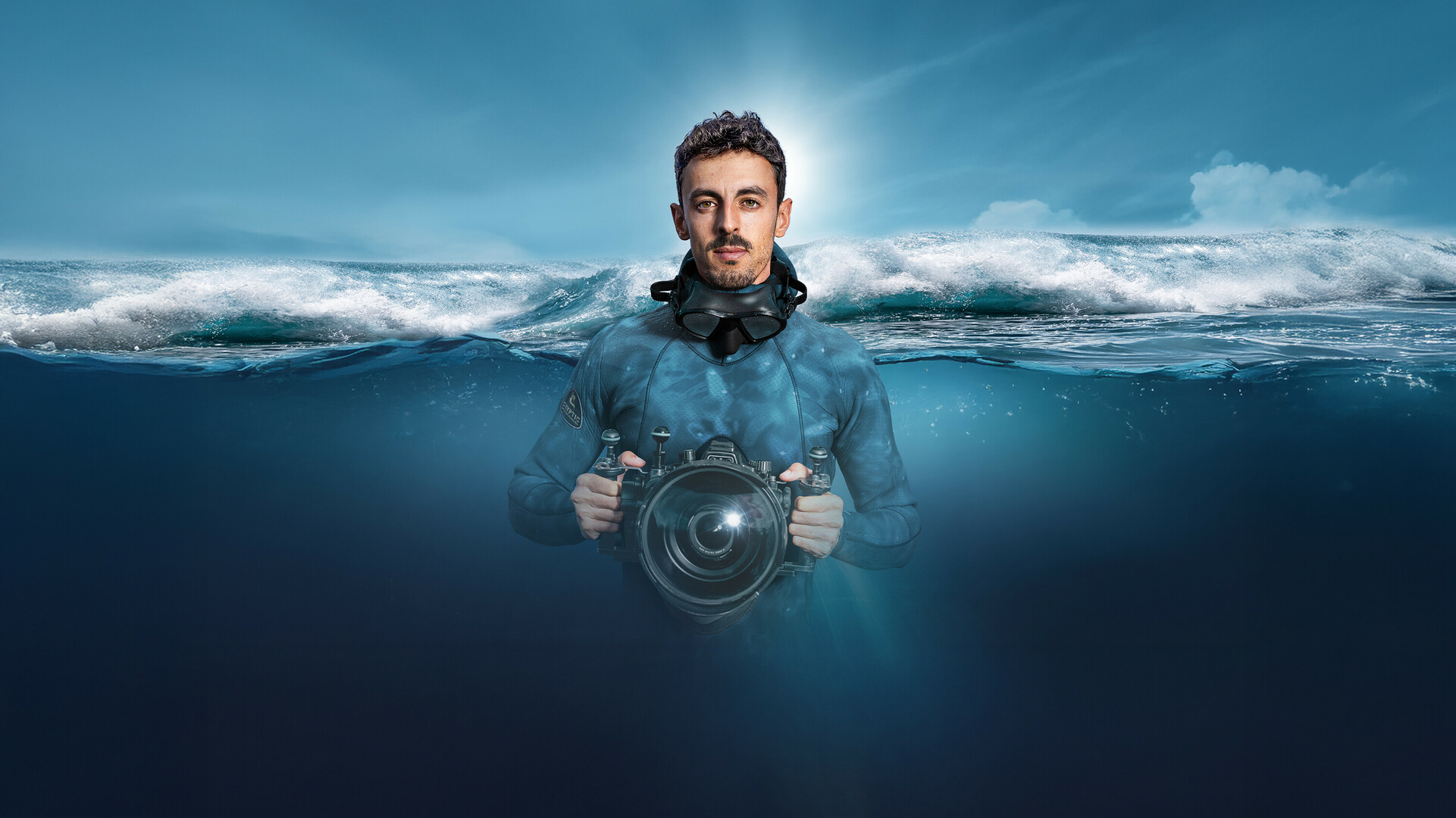 Diver with a diving mask and underwater camera in the ocean, half-submerged, with a breaking wave in the background.